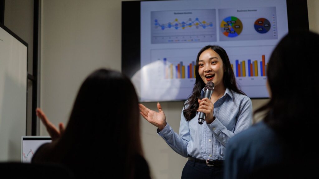 A woman holding a microphone presents business charts to an audience, with animation for presentation bringing the graphs and data visualizations to life on a large screen behind her.