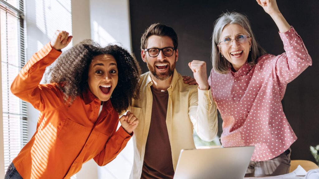 Three people standing indoors by a laptop, smiling and raising their fists in celebration—perhaps they've just received exciting news about Nonprofit Animation Grants.