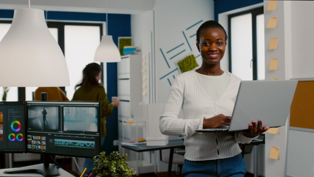 A woman holding a laptop stands and smiles in a modern office, where people work and a computer displays impactful animation for branding strategies.