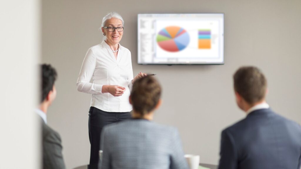 A woman in business attire uses Animation for Presentation as she presents data on a screen with charts to three seated colleagues in a conference room.