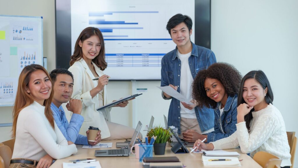 Seven people sit and stand around a table with laptops, papers, and coffee, smiling at the camera. A screen behind them displays bar graphs, charts, and an animation for presentation in the background.