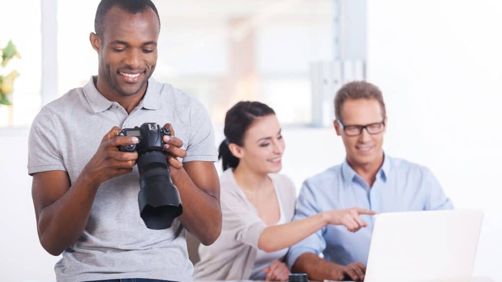 A man holding a camera stands in the foreground, while a woman and a man sit at a desk looking at a laptop, discussing storytelling techniques for content marketing and pointing at the screen.