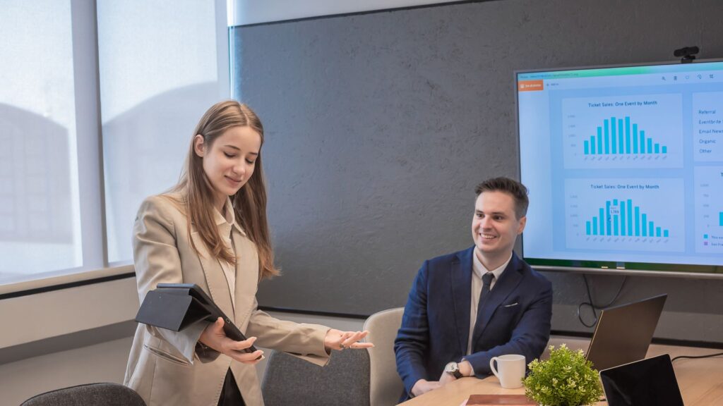 A woman presents data on a tablet to a man in a business meeting room, with charts and animation for presentation displayed on a screen in the background.