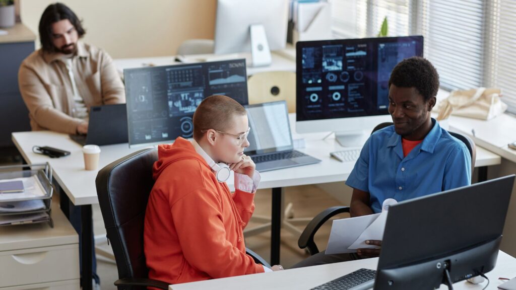 Three people work in a modern UK office with computers; two discuss pricing at a desk while one works alone. Screens display charts, data, and details about 30 second animation cost.