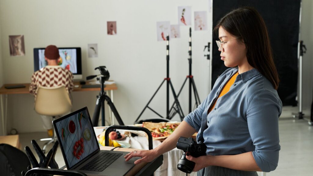 A woman holding a camera looks at a laptop screen in a photography studio, possibly checking out an Animation Pricing UK guide; another person works at a computer in the background.