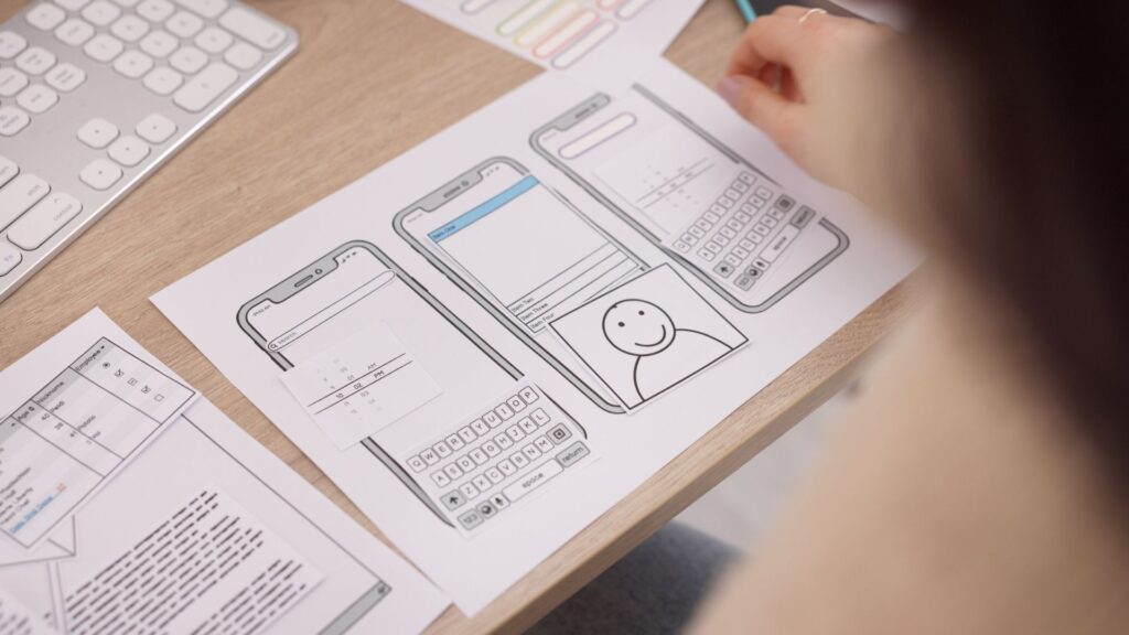 A person reviews printed smartphone wireframes and design sketches on a desk, considering animation for mobile app features, alongside a keyboard and other design documents.
