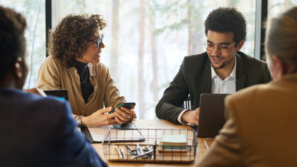 Four people sit around a table in a meeting room, two of them discussing 30 Second Animation Cost UK while using a smartphone and a laptop, with office supplies on the table.