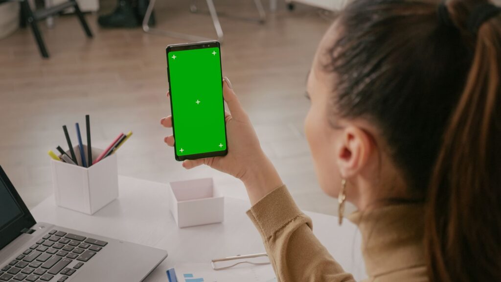 A woman sitting at a desk holds up a smartphone with a green screen, perfect for showcasing an Animation for Mobile App. A laptop, pens, and office supplies are visible on the desk.