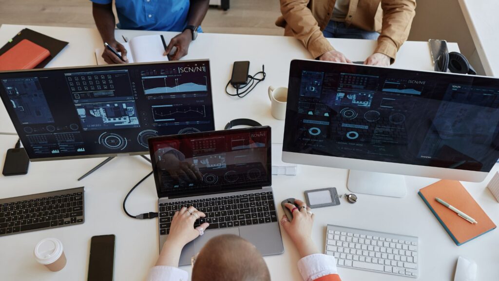 People work at a desk with multiple monitors displaying data and charts—some review an animation value guide, one writes notes on 30 second animation cost UK, while tech devices and office supplies fill the table.