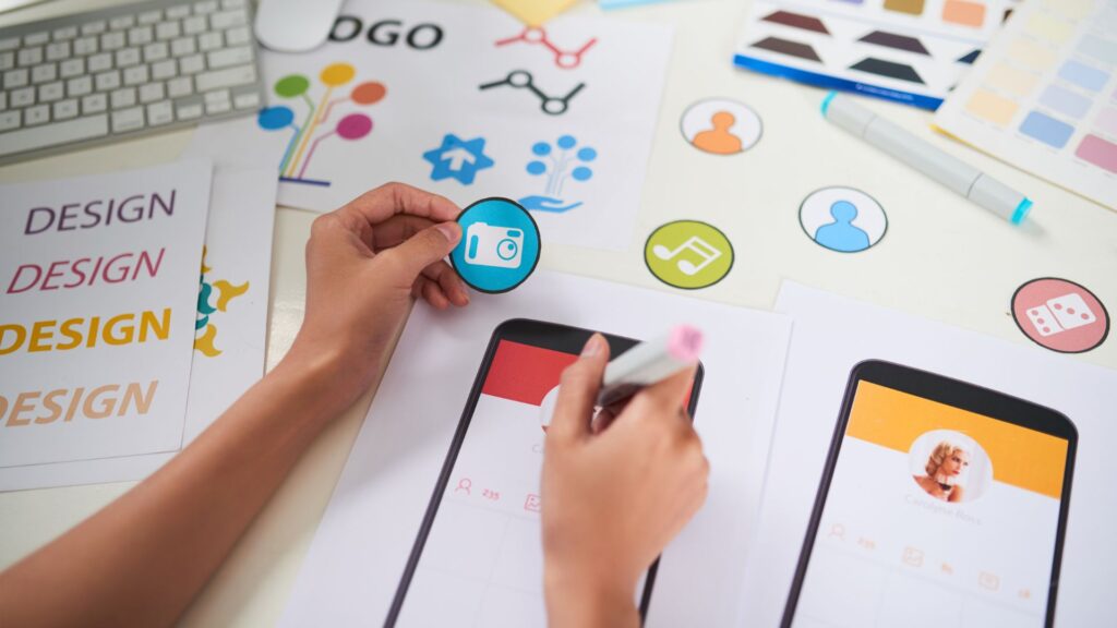 Person designs a mobile app interface with paper mockups, icon cutouts, and color swatches on a desk, planning smooth animation for mobile app transitions.