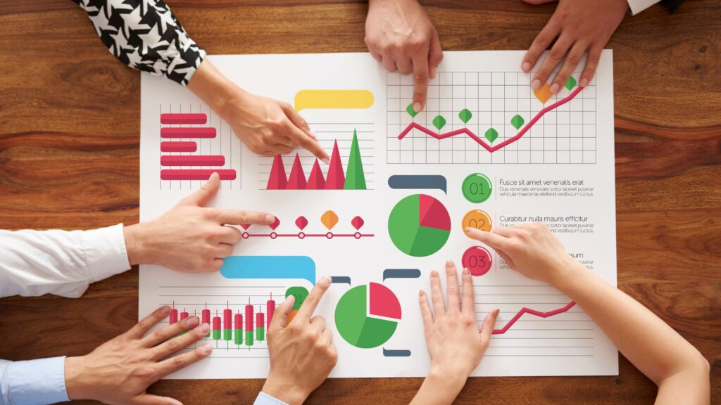 Several hands point to colorful charts and graphs on a large sheet of paper on a wooden table, suggesting a collaborative discussion about animation pricing UK or reviewing an animation value guide.