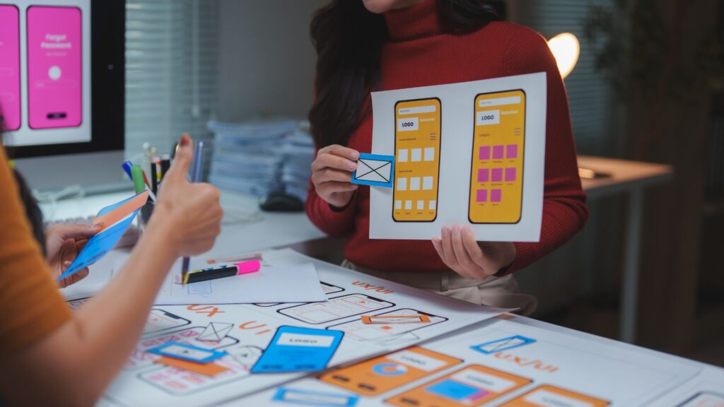 Two people discussing mobile app interface designs, reviewing printed wireframes and mockups, while also considering animation for mobile app features among the design materials and sketches spread out on the table.