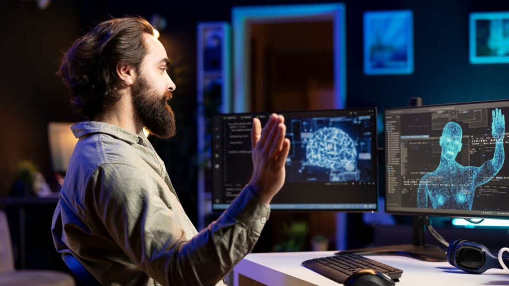 A man sits at a desk waving to a screen, with multiple monitors displaying digital brain graphics and code in a dimly lit room, embodying animation breakthroughs in creative technology.